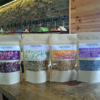 Pacotes de flores secas de rosa, camomila, calêndula e lavanda em pacotes de papel reciclado com etiquetas coloridas sobre mesa de madeira.