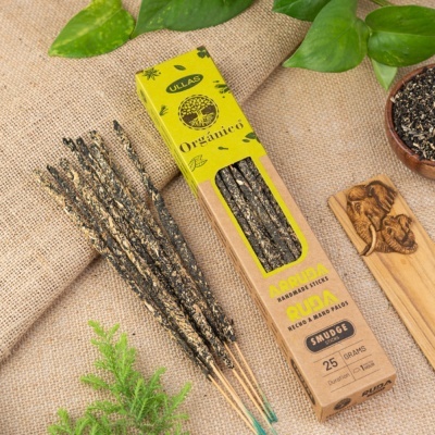 Paus de defumação artesanais embalados em caixa de papel reciclado amarela com texto e decoração natural sobre tecido de juta.