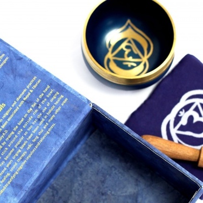 Conjunto com taça tibetana dourada, bastão de madeira, almofada azul com símbolo branco e caixa azul com texto