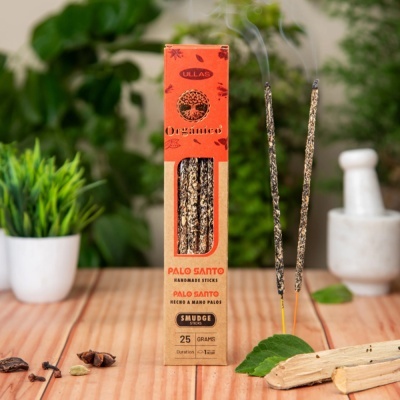 Caixa de incenso Palo Santo Orgânico com dois pauzinhos acesos e folhas verdes sobre mesa de madeira