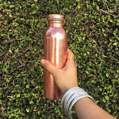 Garrafa de cobre com tampa segurada por mão com pulseiras prateadas e fundo de plantas