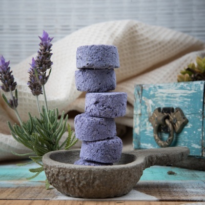 Cinco pastilhas de banho roxas empilhadas numa colher de madeira com lavanda e caixa azul ao fundo
