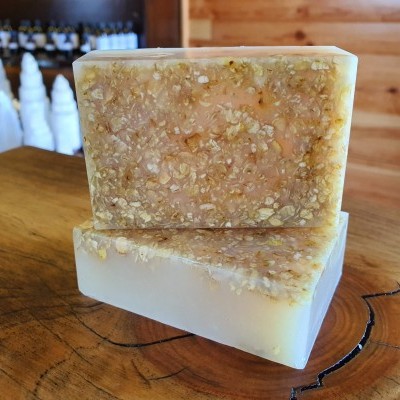 Dois sabonetes artesanais retangulares com flocos cor de bege claro na parte superior, sobre superfície de madeira