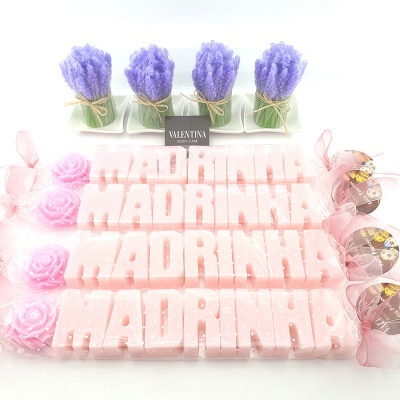 Velas rosas com texto 'MADRINHA', decoradas com rosas e laços, sobre mesa branca com decoração com ramos roxos.