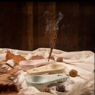 Incensários cerâmicos em tons pastel com incenso aceso, folhas secas e decoração sobre tecido branco