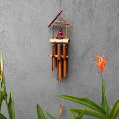 Sino dos ventos em bambu com figura de Buda pendurado numa parede cinza com plantas verdes e flor laranja