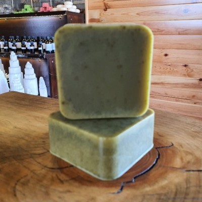 Dois sabonetes quadrados verdes sobre superfície de madeira rústica, com prateleiras de garrafas ao fundo.