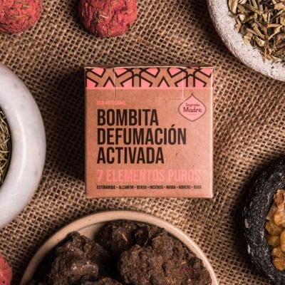 Caixa rosa de Bombita Defumación Activada cercada por tigelas com ervas e resinas em fundo de tecido rústico.