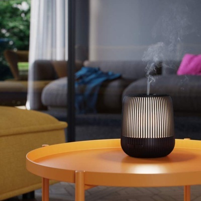 Difusor preto com riscas claras numa mesa laranja em sala com sofá cinzento e almofada rosa