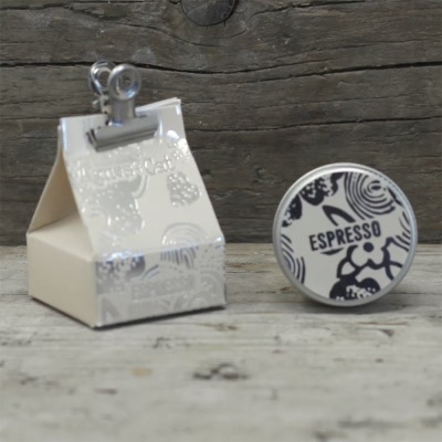 Caixa pequena creme com tampa prateada e clip com caixa redonda preta e branca rotulada como ESPRESSO em fundo de madeira