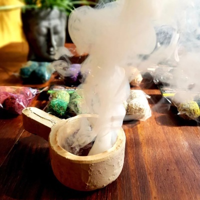 Incenso aceso em suporte de cerâmica bege com sacos coloridos ao fundo e estátua desfocada