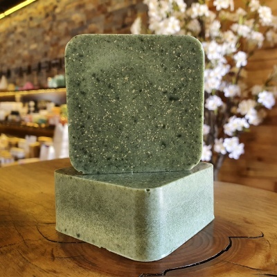 Sabonete cúbico verde com textura granulada sobre mesa de madeira, fundo com flores brancas e prateleiras