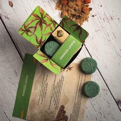 Produto cosmético verde com folhas, pastilhas verdes e barra dourada 'ENERGIA LIMPIA' em caixa aberta sobre mesa de madeira.