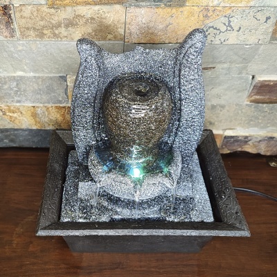 Fonte decorativa cinza com esfera central iluminada e base quadrada sobre mesa de madeira