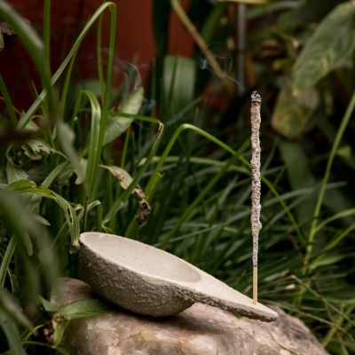 Incensário cinza claro texturizado com incenso aceso em pedra rodeado por plantas verdes