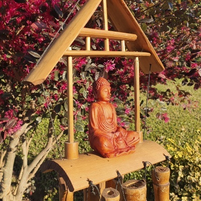 Escultura de Buda em madeira com estrutura de madeira e sinos de vento, em ambiente exterior com flores.