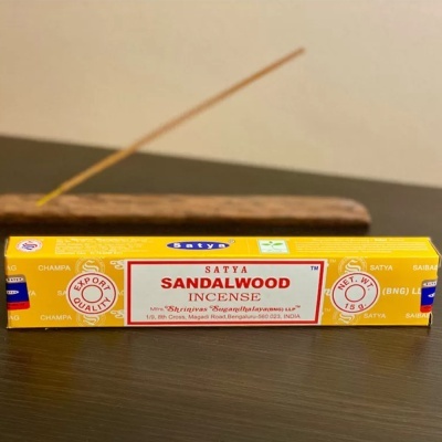 Caixa amarela de incenso Satya Sandalwood sobre mesa escura com incenso aceso em suporte de madeira ao fundo