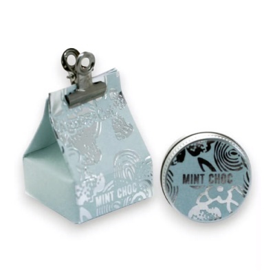 Saco de papel azul claro com padrão branco e texto MINT CHOC, fechado com clipe metálico, ao lado de lata redonda com mesma cor, padrão e texto.