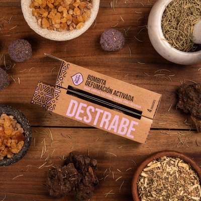 Caixa de bombitas de defumação activada com texto DESTRABE, cercada por resinas, ervas secas e madeira sobre madeira escura