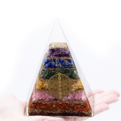 Pirâmide decorativa com camadas coloridas de pedras e símbolo dourado no centro, segurada por uma mão.