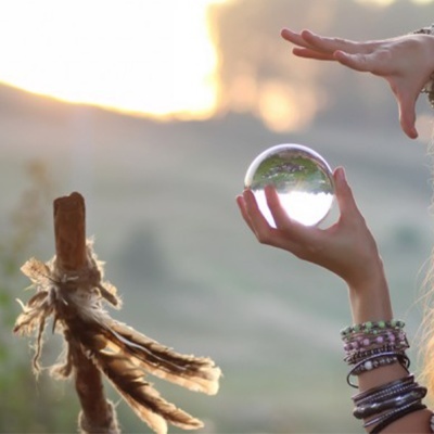 Mãos segurando esfera de cristal próxima a um objeto de madeira com penas ao pôr do sol