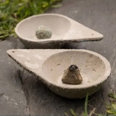 Dois suportes para incenso triangulares cinza com cones de incenso, sobre superfície escura ao ar livre.