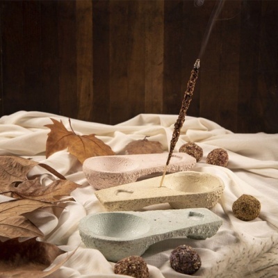 Três incensários de cerâmica em tons pastel com incenso aceso, folhas secas e bolinhas decorativas sobre tecido branco