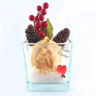 Decoração natalícia em vaso de vidro com pinhas, bagas vermelhas, cena em marfim e texto Feliz Natal