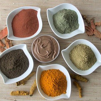 Seis tigelas brancas com pós coloridos e pasta castanha, sobre superfície bege com raízes secas