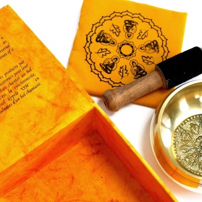 Conjunto de taça tibetana dourada, malho de madeira, pano amarelo com desenhos e caixa amarela com texto.