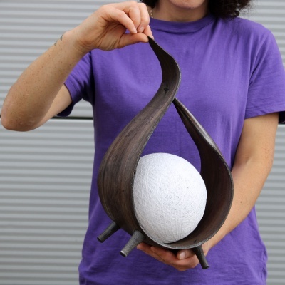 Objeto decorativo em madeira escura com esfera branca, segurado por pessoa com t-shirt roxa