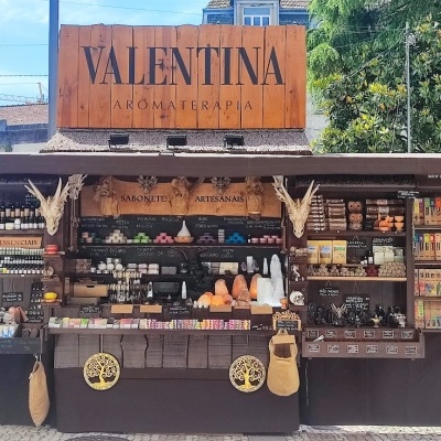 Banca de madeira ao ar livre com produtos artesanais e cartaz VALENTINA A LOJA DA PRAÇA