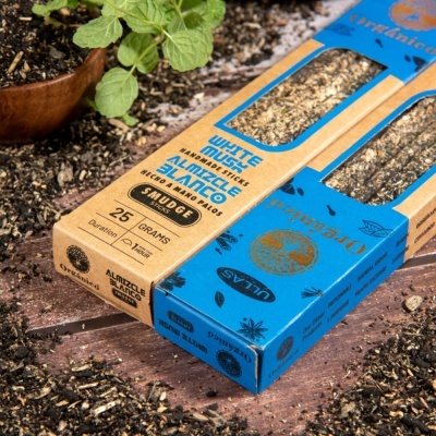 Caixa de incenso White Musk Almitzcle Blanco em embalagem castanha e azul, sobre terra com planta verde.