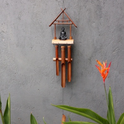 Sino dos ventos de bambu com estátua de Buda pendurado numa parede cinza com plantas verdes e flor laranja.