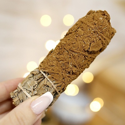 Incenso de sálvia bruxa amarrado com fio branco, com cobertura marrom, segurado por mão com unhas pintadas de rosa claro