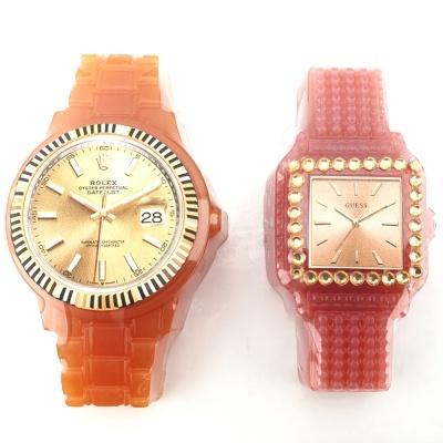 Dois relógios de pulso, um Rolex dourado e um Guess rosa, com braceletes de plástico.