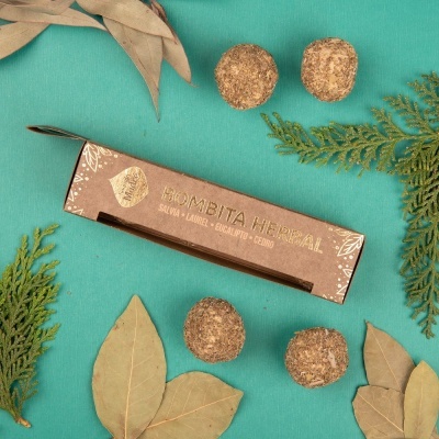 Caixa de bombitas herbais com folhas secas e bolas marrons sobre fundo verde turquesa