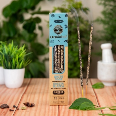 Caixa de incensos orgânicos de sálvia branca com dois bastões acesos e folhas verdes sobre mesa de madeira.