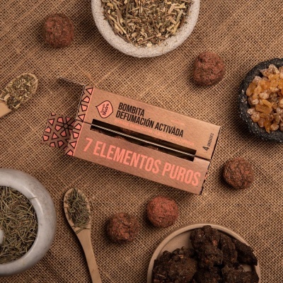 Embalagem de papel castanha com texto 'BOMBITA DEFUMAÇÃO ACTIVADA 7 ELEMENTOS PUROS' sobre tecido rústico com elementos naturais ao redor