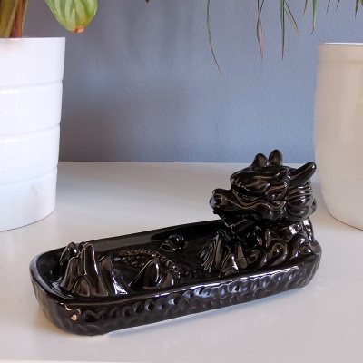 Incensário de cerâmica preto em forma de barco com dragão, sobre superfície branca entre vasos brancos.