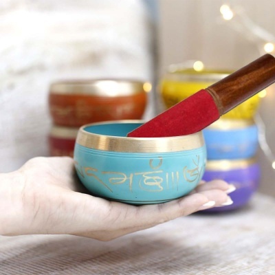 Tigela azul turquesa com padrão dourado e bastão vermelho em mão, com outras tigelas coloridas ao fundo