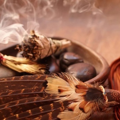 Leque de penas castanhas, ramo de ervas a arder e pedras negras numa superfície de madeira