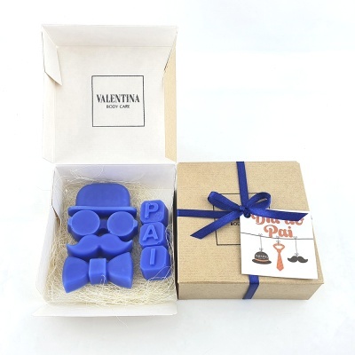 Sabonetes azuis em forma de óculos, bigode, laço e letras PAI numa caixa branca aberta ao lado de caixa bege fechada com fita azul e etiqueta ótiomo pai.