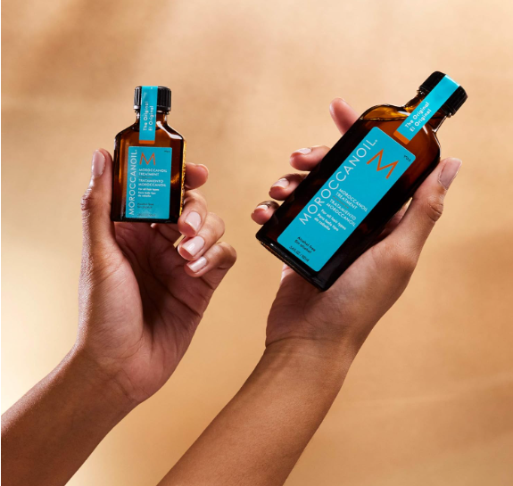 Mãos segurando frascos de óleo Moroccanoil em vidro castanho com rótulos azuis.