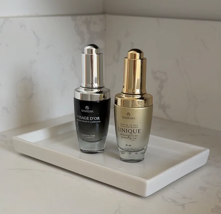 Dois frascos de sérum, um preto prateado e outro dourado, numa bandeja branca sobre bancada branca