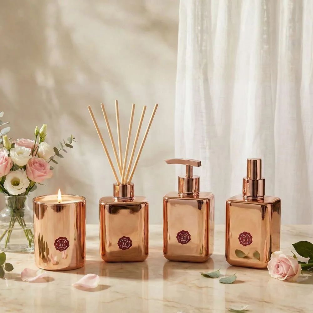 Conjunto decorativo em vidro bronze com vela, difusor, dispenser e spray junto a flores e pétalas numa superfície marmoreada