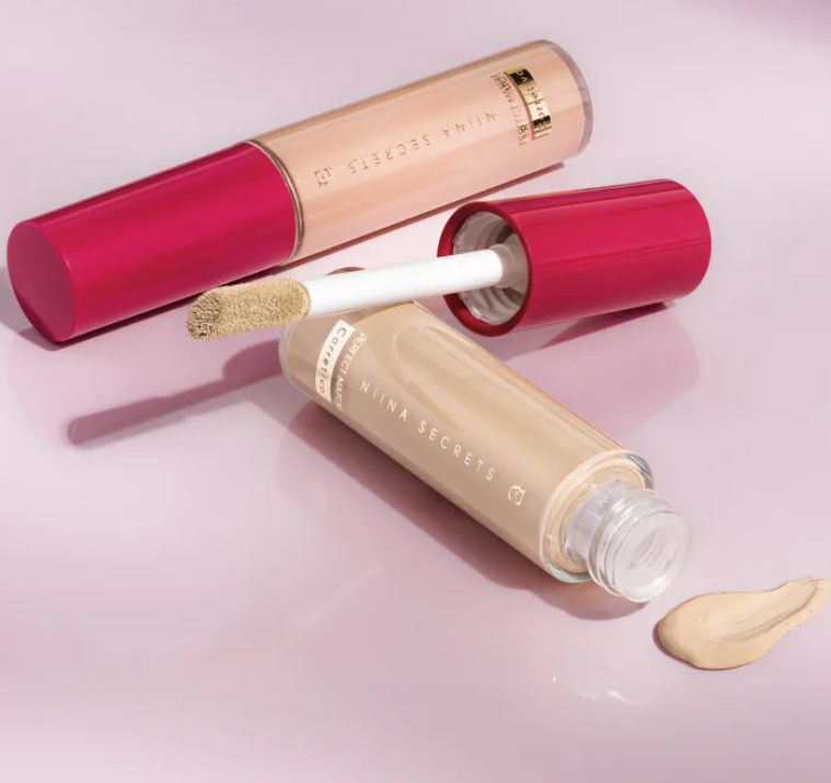 Dois tubos de corretor líquido bege claro com tampa vermelha e aplicador, da marca NINA SECRETS, em fundo branco