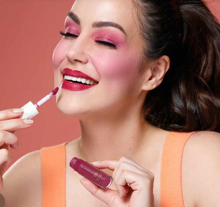 Mulher com maquilhagem rosa a aplicar batom líquido vinho num fundo coral