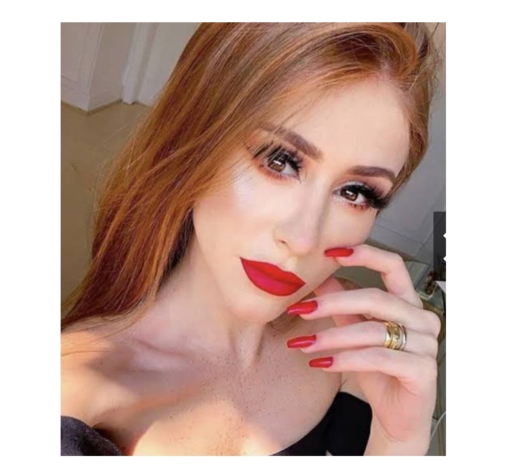 Mulher com cabelo laranja-avermelhado, maquilhagem vermelha, unhas vermelhas, anel dourado e vestido preto