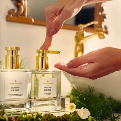 Dois dispensadores de vidro quadrados dourados com cremes e líquides, flores, e mão a pressionar um dispensador.